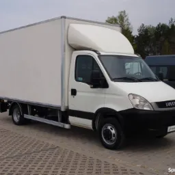 Biały samochód dostawczy Iveco z zabudową kontenerową na kostce brukowej, widoczny w tle inny pojazd i znak drogowy.