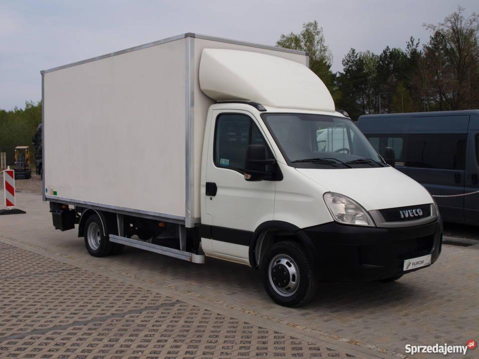 Biały samochód dostawczy Iveco z zabudową kontenerową na kostce brukowej, widoczny w tle inny pojazd i znak drogowy.