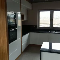 Nowoczesna kuchnia z białymi, lakierowanymi frontami szafek, czarnym blatem i drewnianymi elementami wykończeniowymi; widoczny piekarnik w zabudowie i wyspa kuchenna z czarnym blatem.