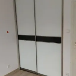 Dwudrzwiowa szafa przesuwna z białymi frontami, poziomym czarnym pasem i metalową ramą w jasnym wnętrzu, widoczna instalacja elektryczna na suficie.