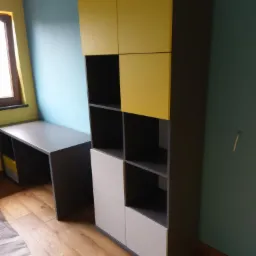 Regał z szafkami i otwartymi półkami w kolorach żółtym i szarym, dopasowany do biurka w tym samym stylu, ustawiony na drewnianej podłodze w pokoju z niebiesko-zielonymi ścianami.