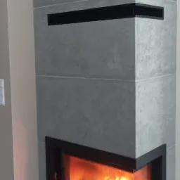 Nowoczesny kominek narożny z szarą obudową imitującą beton, z czarnymi wstawkami i paleniskiem z widocznym ogniem, umieszczony w rogu pomieszczenia.