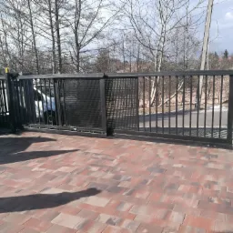 Dwudzielna brama przesuwna z metalową konstrukcją i ażurowym wypełnieniem, zamontowana na posesji z kostki brukowej w odcieniach czerwieni i szarości, widoczna z perspektywy lekko z dołu,...