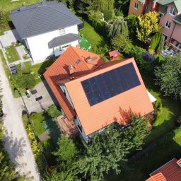 SOLAR SQUARE SP. Z O.O. - Widok z drona na dom z pomarańczową dachówką i panelami fotowoltaicznymi na dachu, otoczony zielenią i innymi budynkami.