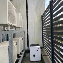 Zewnętrzne jednostki klimatyzacyjne na metalowej konstrukcji, obok biała obudowa z logo SMA, widok zza szarego płotu, ściana budynku z widocznymi śladami wilgoci.