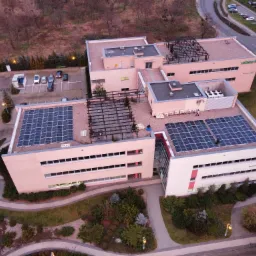 Widok z góry na budynek z panelami słonecznymi na dachu, otoczony zielenią i parkingiem, z widoczną ulicą i drzewami w tle, sfotografowany o zmierzchu.