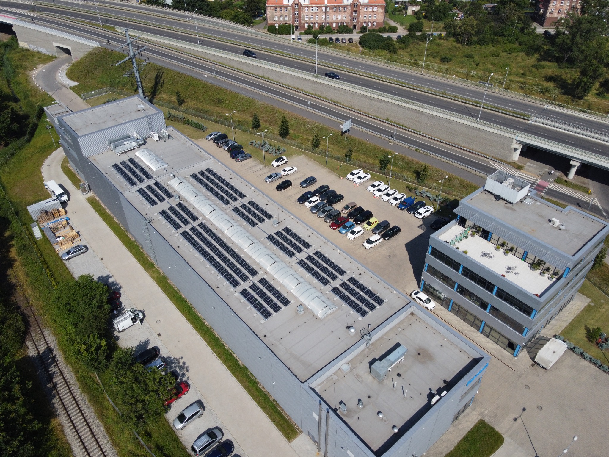 Widok z lotu ptaka na budynek z panelami słonecznymi na dachu, przylegający parking z samochodami, obok budynek biurowy i autostrada w tle.