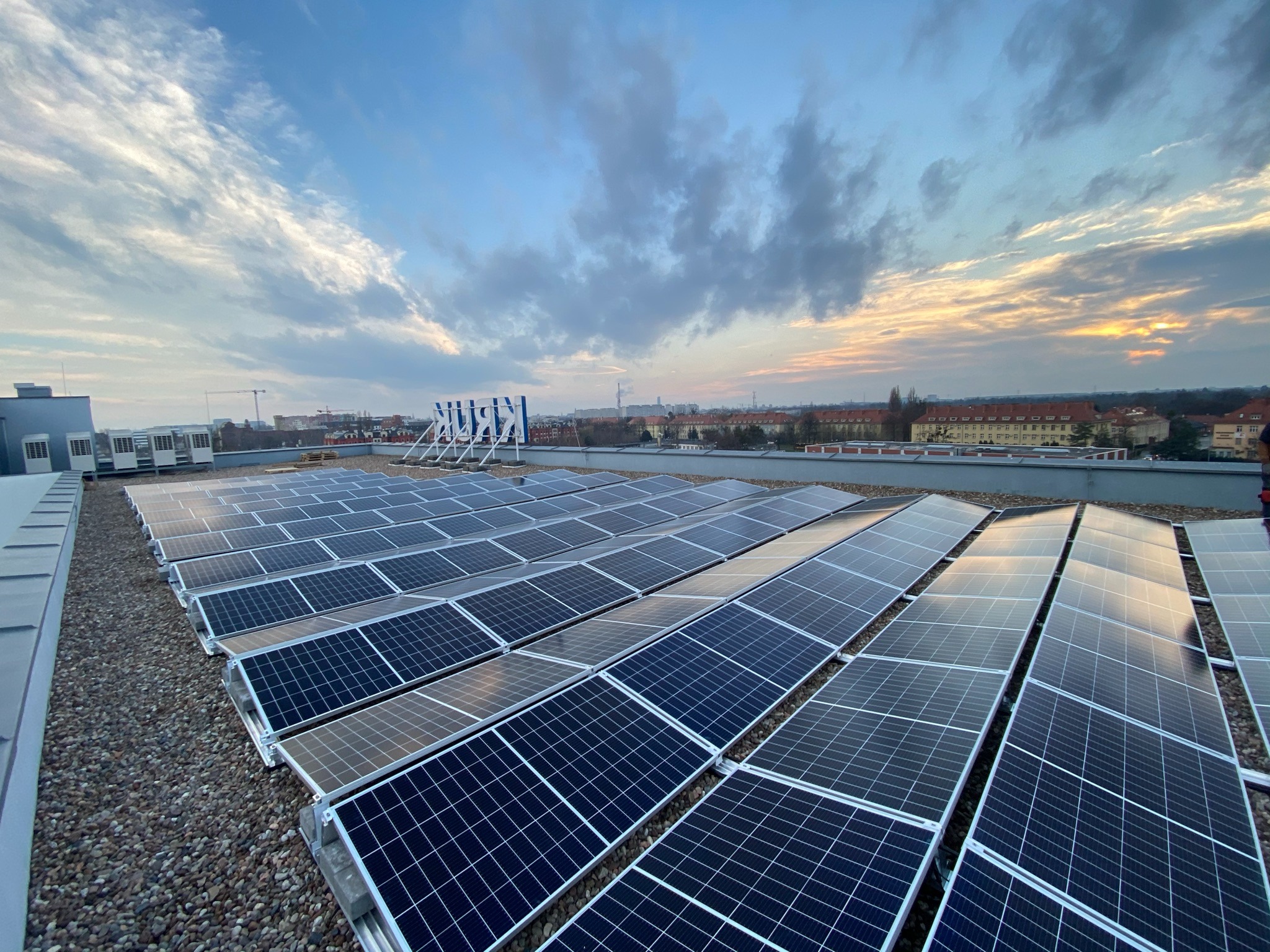 Rząd paneli słonecznych na płaskim dachu pokrytym żwirem, z widokiem na zabudowę miejską i zachodzące słońce na horyzoncie.