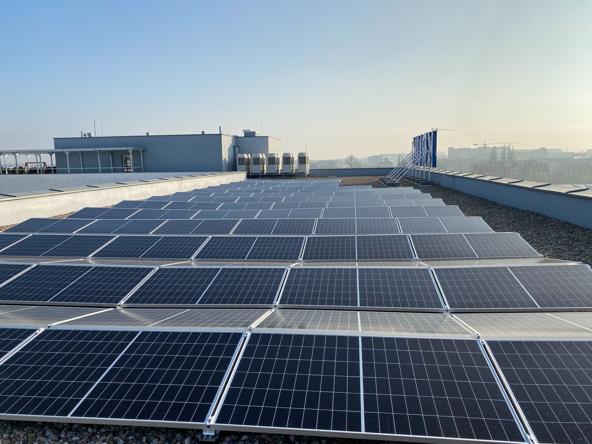 Rząd paneli słonecznych na płaskim dachu budynku z widokiem na miasto w tle, panele ułożone równolegle na żwirowym podłożu, widoczne elementy konstrukcyjne dachu i klimatyzatory.