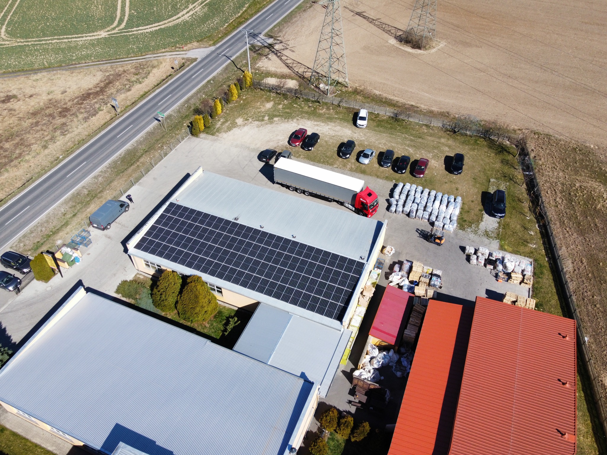 Widok z drona na budynek przemysłowy z panelami słonecznymi na dachu, zaparkowane samochody, ciężarówkę z naczepą, palety z materiałami budowlanymi i wózek widłowy.