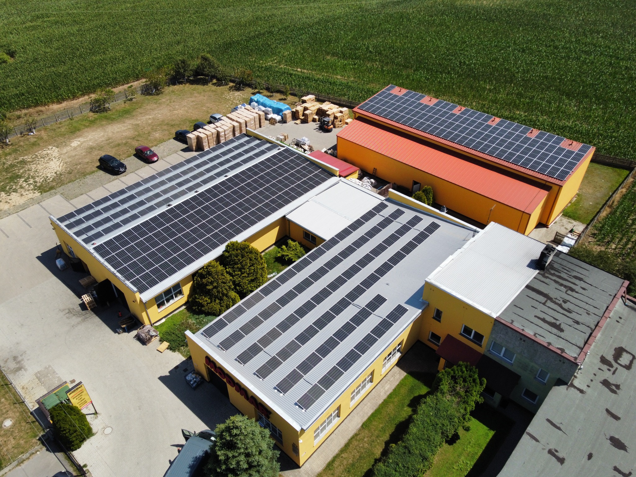 Widok z lotu ptaka na budynki przemysłowe z panelami słonecznymi na dachach, obok składowane palety z materiałami, w tle pole uprawne.