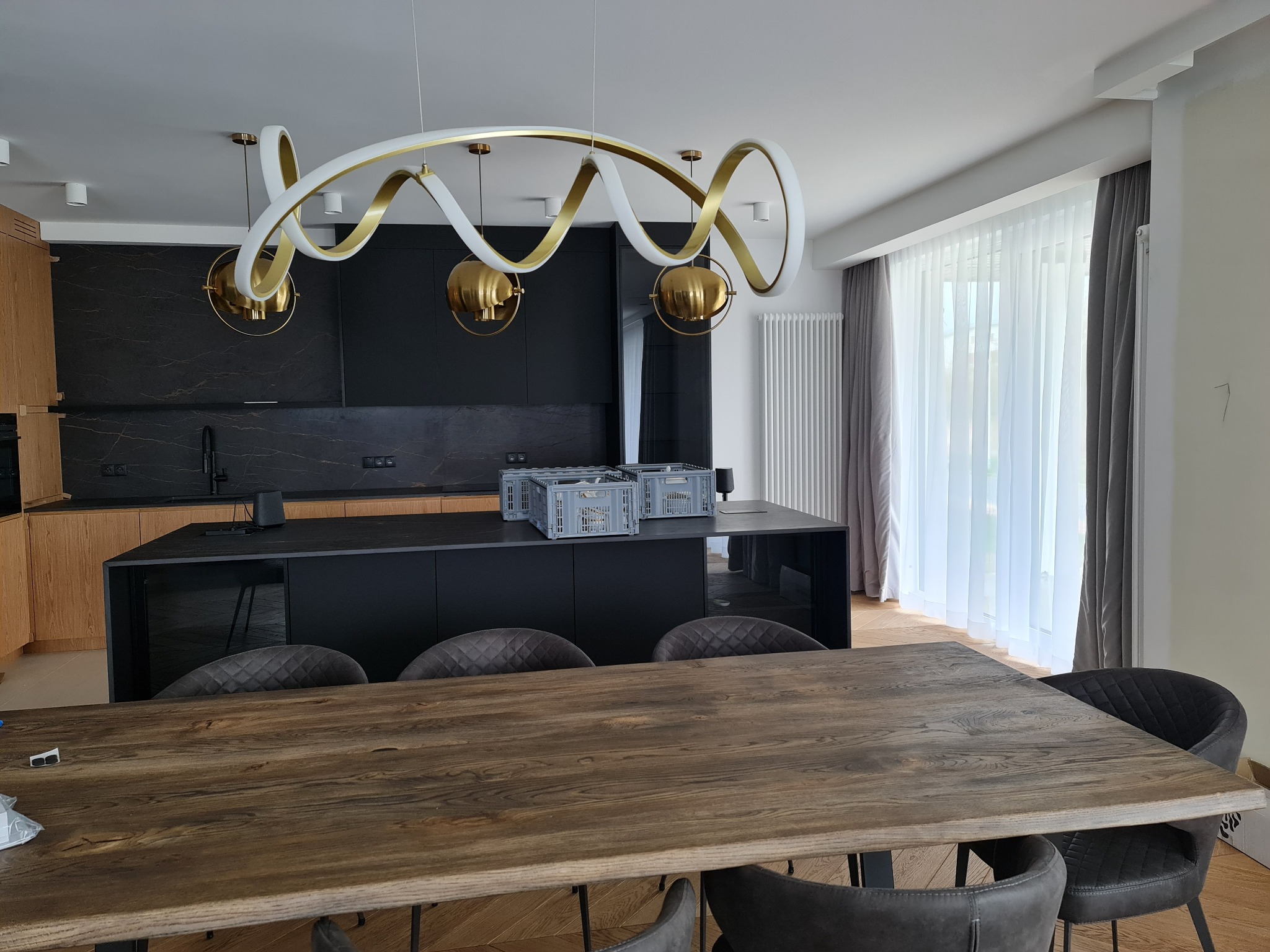 Nowoczesna kuchnia z czarnymi szafkami i wyspą, drewniany stół z szarymi krzesłami, dekoracyjne oświetlenie sufitowe w kształcie spirali, szare plastikowe skrzynki na wyspie, jasne zasłony w oknie.