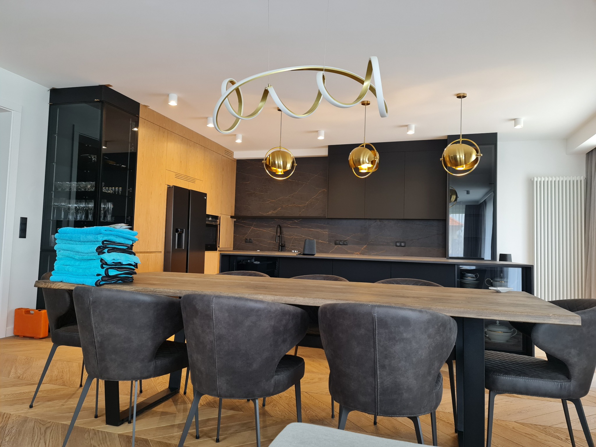 Nowoczesna kuchnia z drewnianymi i czarnymi szafkami, złotymi lampami wiszącymi i dużym stołem z szarymi krzesłami, na którym leżą sterty błękitnych ręczników.