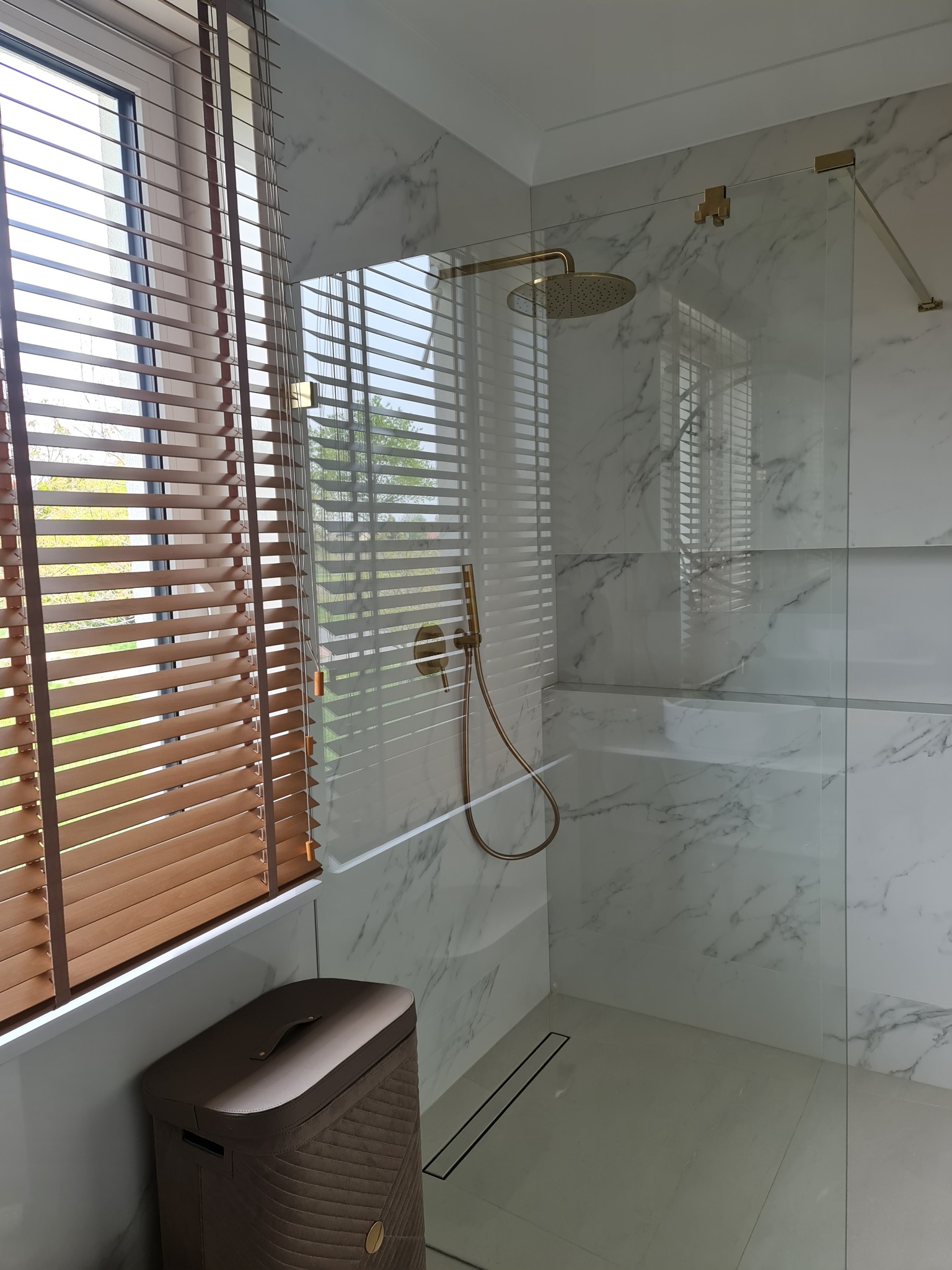 Nowoczesna łazienka z kabiną prysznicową typu walk-in, wykończona płytkami imitującymi marmur, z złotą armaturą i drewnianymi żaluzjami w oknie.