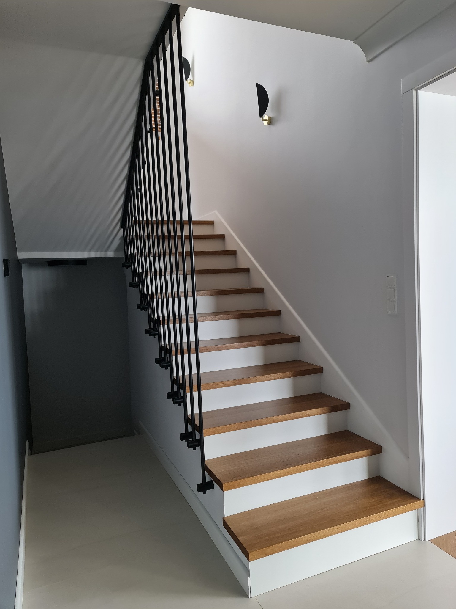 Nowoczesne schody z drewnianymi stopniami i czarną, minimalistyczną balustradą w domu w Markach, oświetlone przez ścienny kinkiet w kształcie półksiężyca.