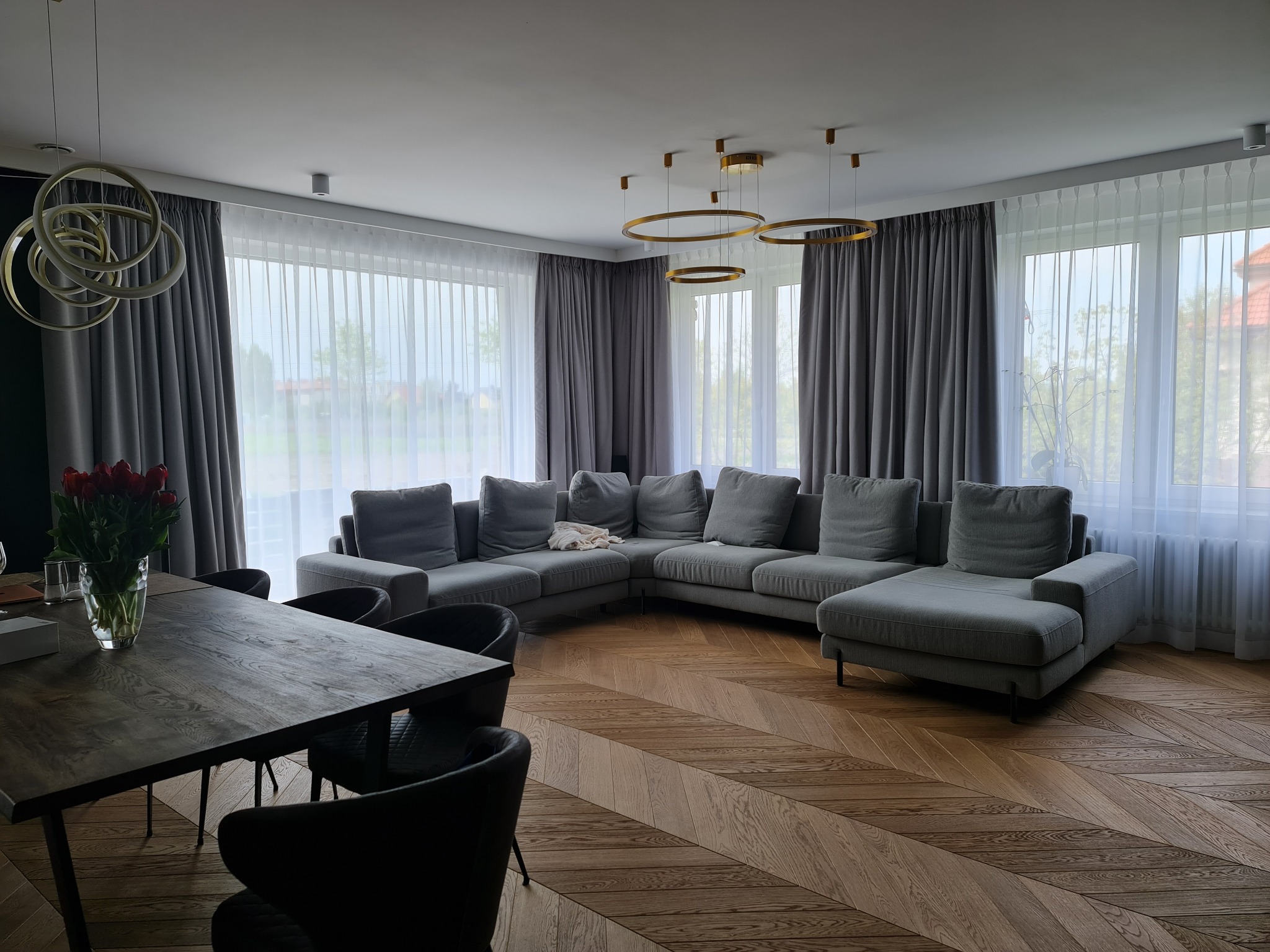 Jasne wnętrze salonu z parkietem w jodełkę, szarą sofą narożną, ciemnym drewnianym stołem z bukietem czerwonych tulipanów i nowoczesnymi lampami pierścieniowymi.