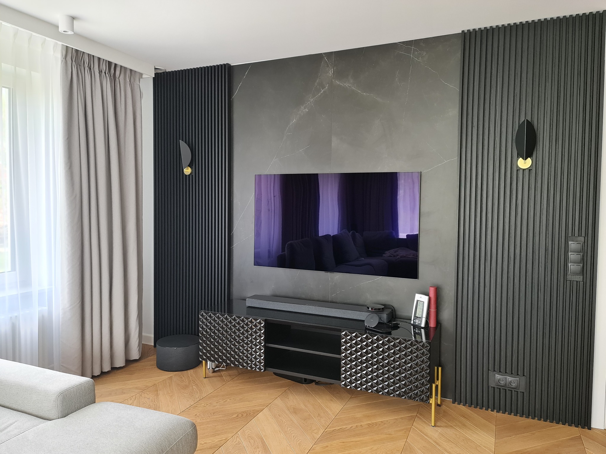 Elegancki salon z marmurową ścianą, panele ścienne w kolorze grafitowym, telewizor, szafka RTV z otwartymi drzwiczkami o geometrycznym wzorze, parkiet w jodełkę i szara sofa.
