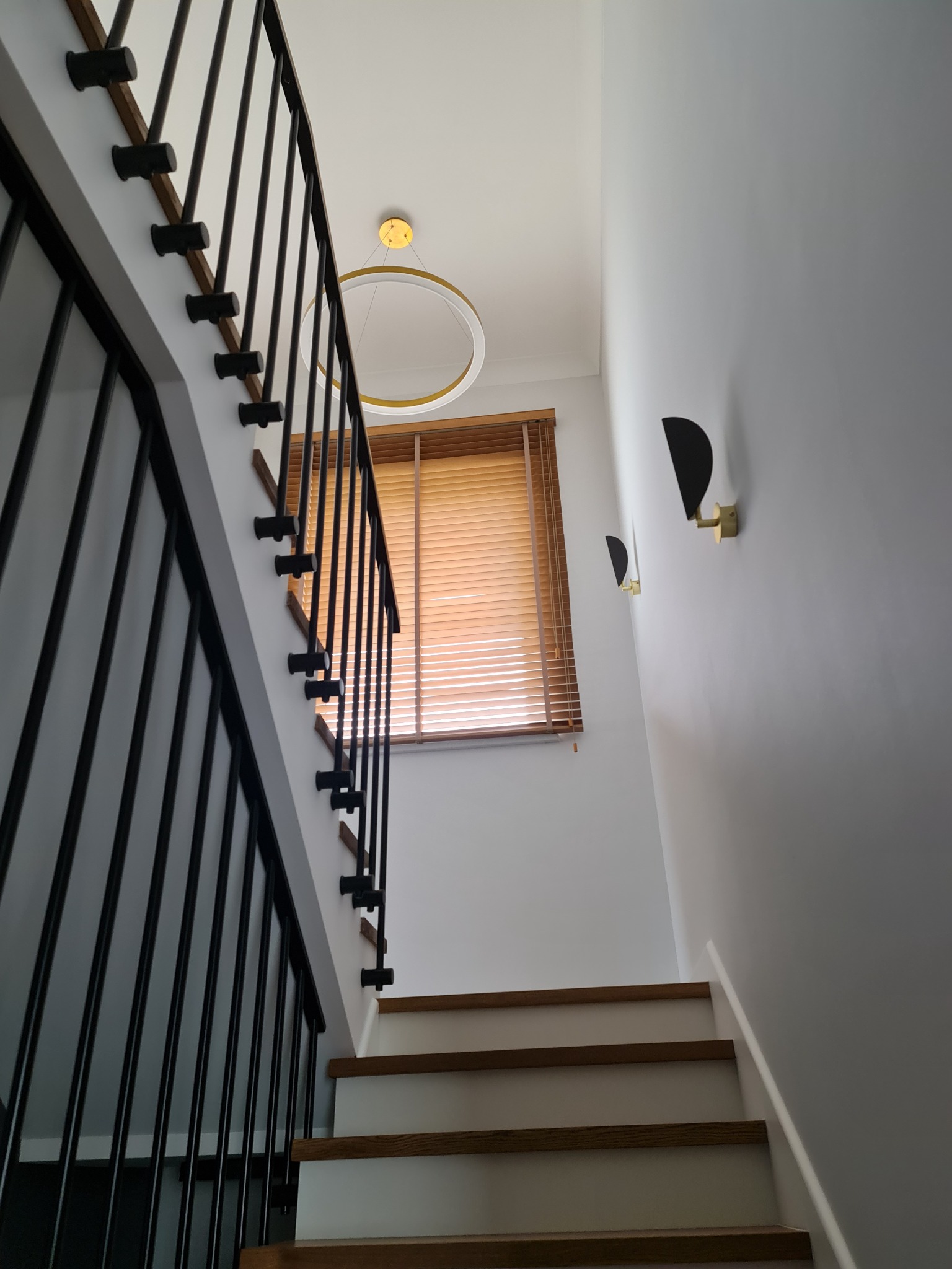 Nowoczesne schody z czarną balustradą i drewnianymi stopniami, oświetlone okrągłą lampą sufitową i dwoma kinkietami w kształcie półksiężyca, widok z dołu.