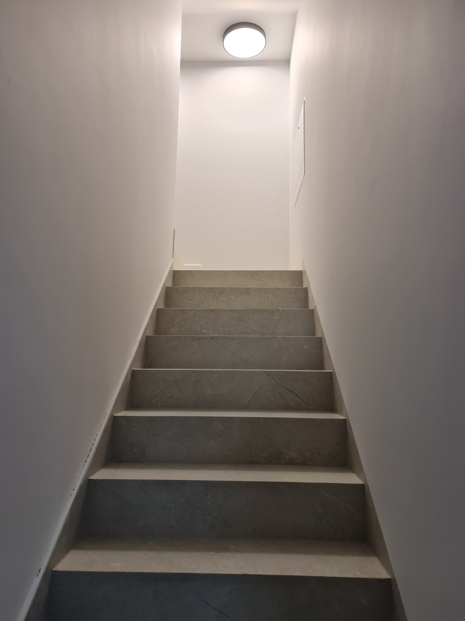 Nowoczesne, szare schody z marmurowym wzorem, oświetlone okrągłą lampą sufitową, prowadzące w górę w minimalistycznym, białym wnętrzu.