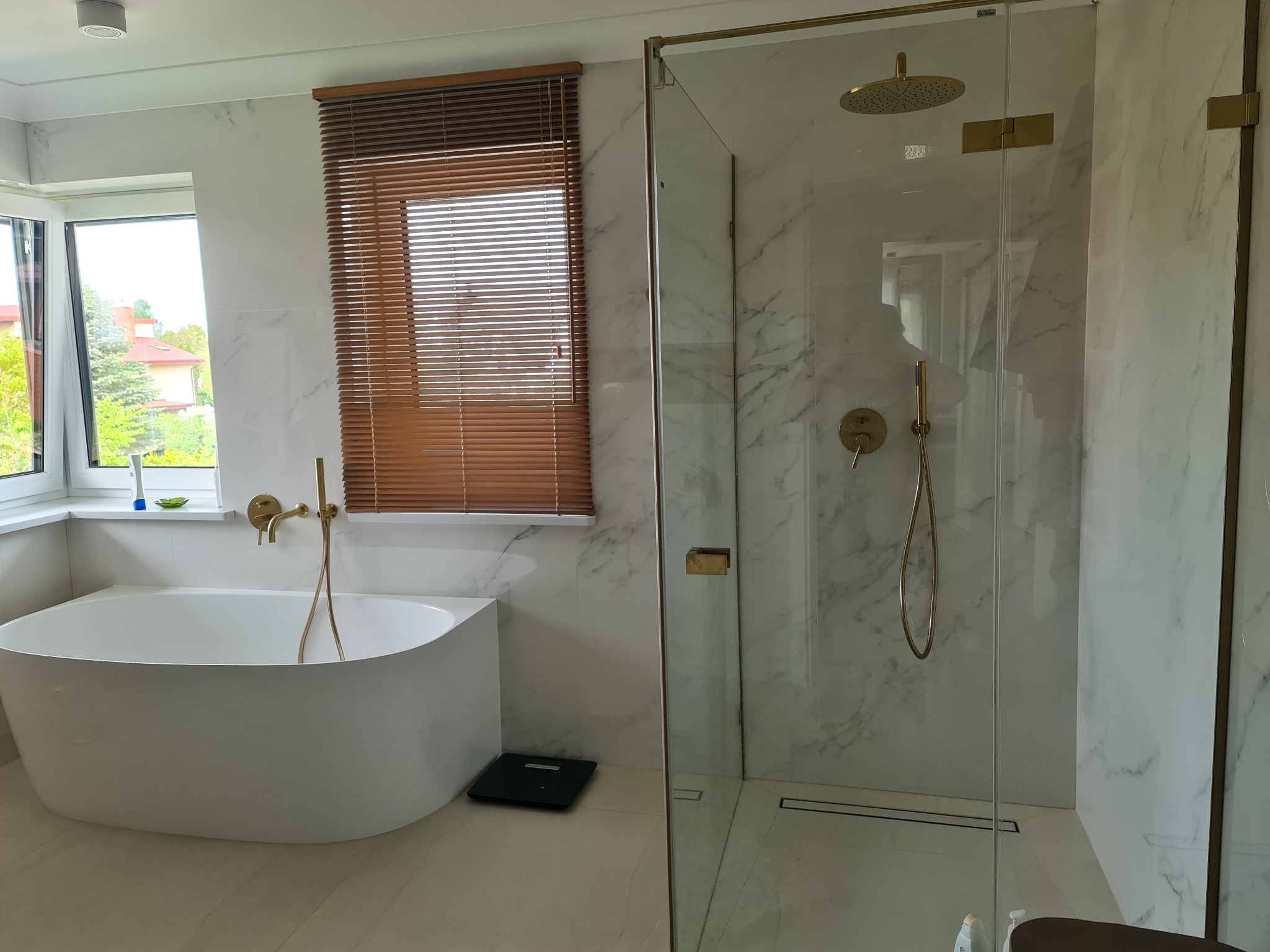 Nowoczesna łazienka z wolnostojącą wanną, kabiną prysznicową typu walk-in z złotą armaturą, marmurowymi płytkami i drewnianymi żaluzjami.