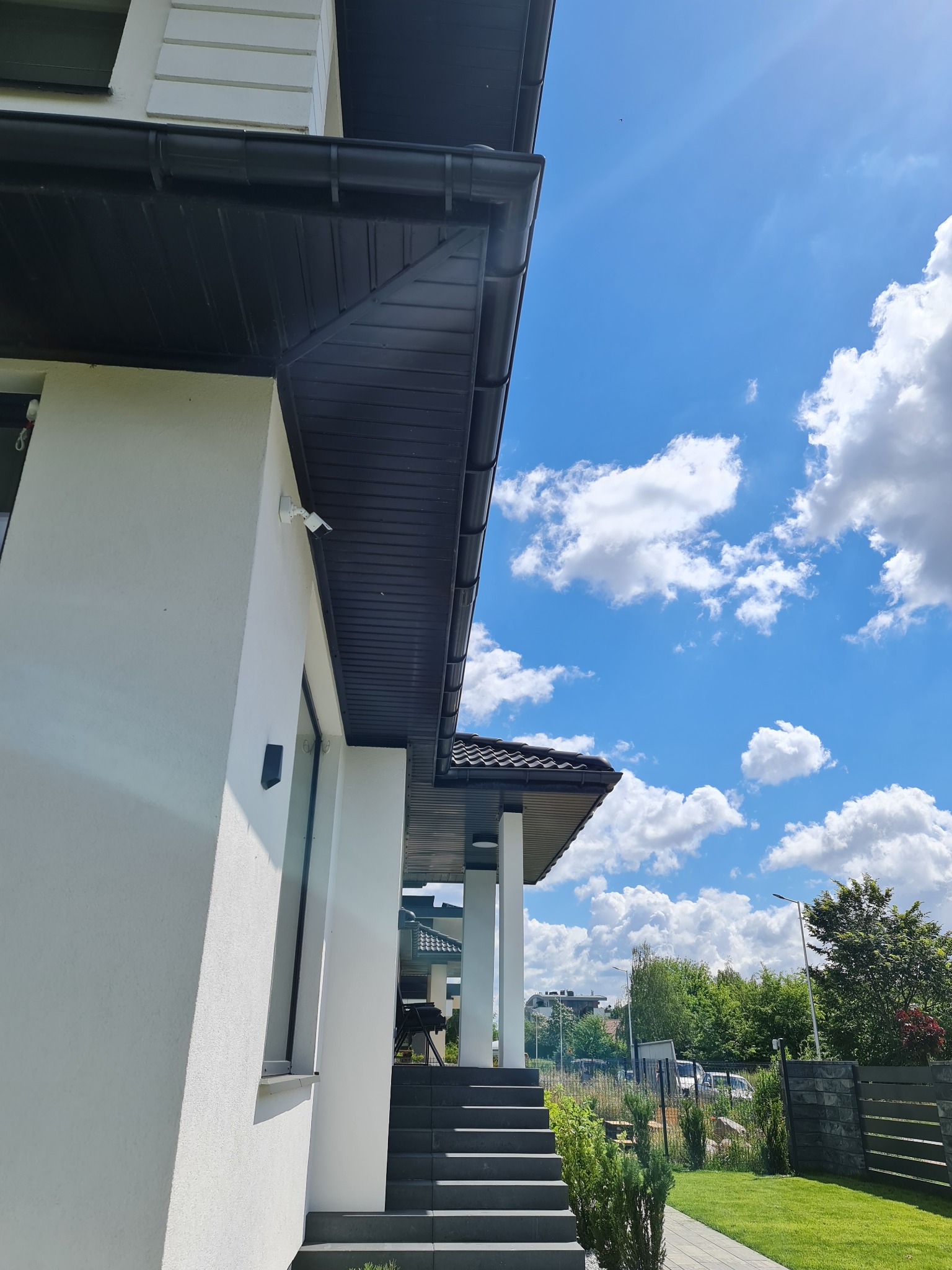 Nowoczesny dom jednorodzinny z białym tynkiem i ciemną podbitką dachową, widok z dołu w słoneczny dzień.