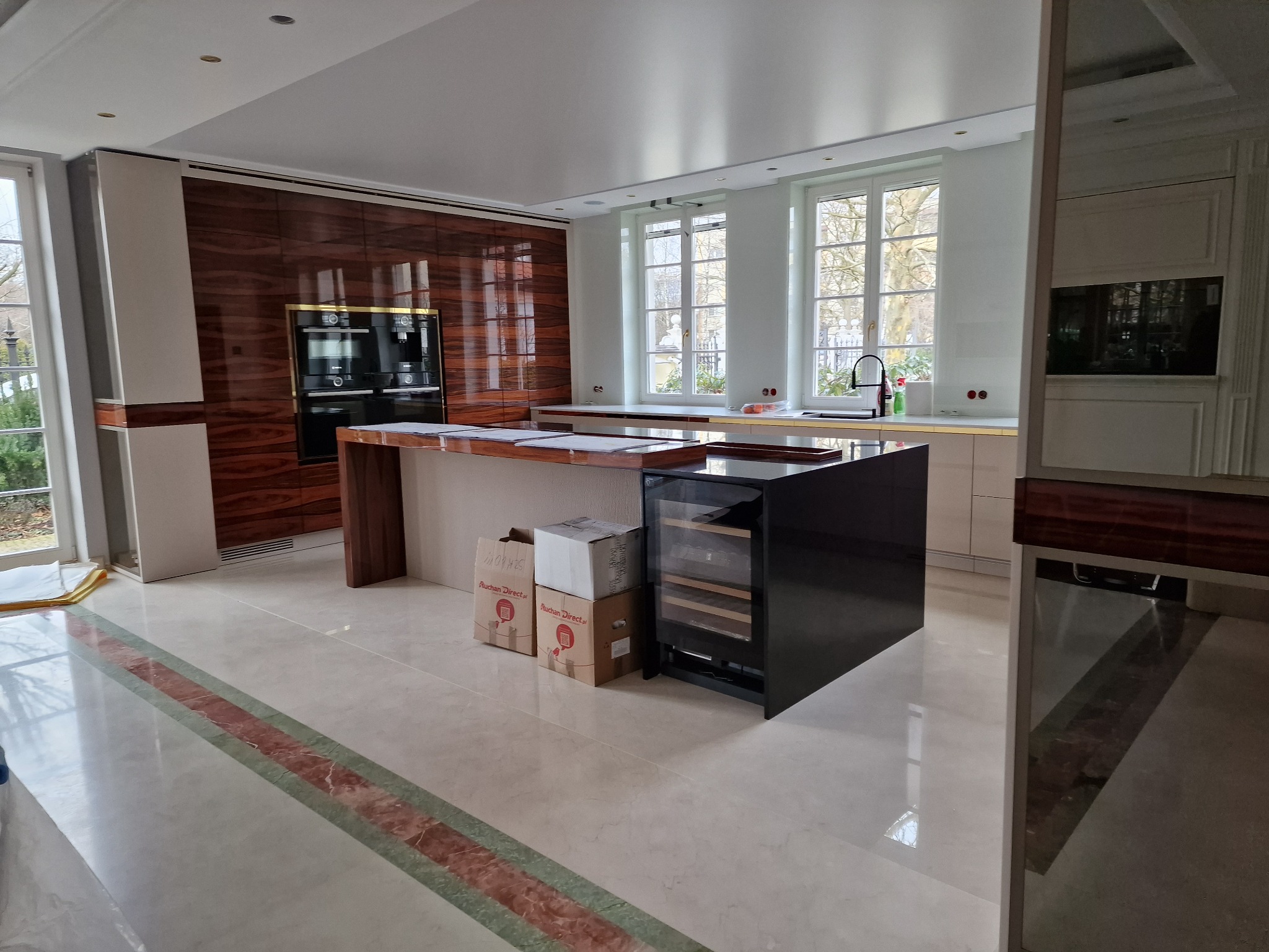 Przestronna kuchnia w trakcie prac wykończeniowych, widoczne zabudowane szafki z forniru, wyspa kuchenna z blatem z ciemnego drewna, podłoga z jasnego marmuru z dekoracyjnym pasem i kartony po...