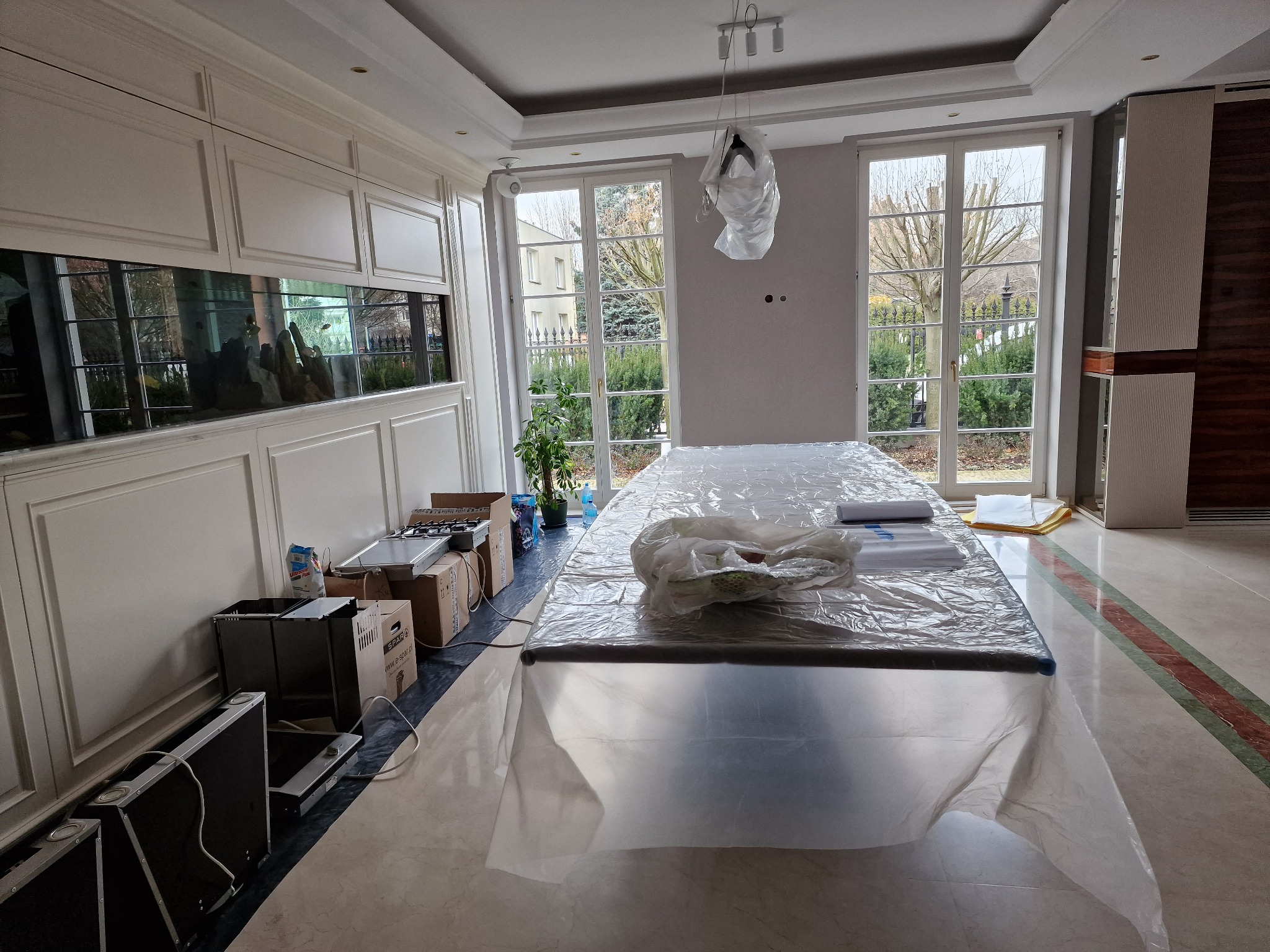 Przygotowania do prac w przestronnym salonie z dużym akwarium: meble i podłoga zabezpieczone folią malarską, widoczne kartony z nowymi urządzeniami kuchennymi.