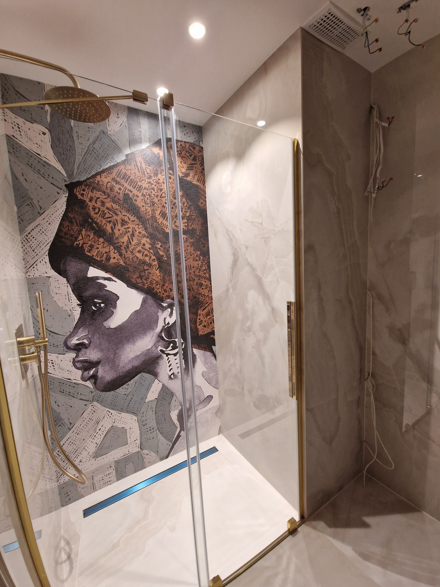 Nowoczesna kabina prysznicowa z grafiką portretu kobiety na szarej ścianie, złote wykończenia armatury i ramy kabiny, widoczne niedokończone podłączenia elektryczne wentylacji sufitowej.