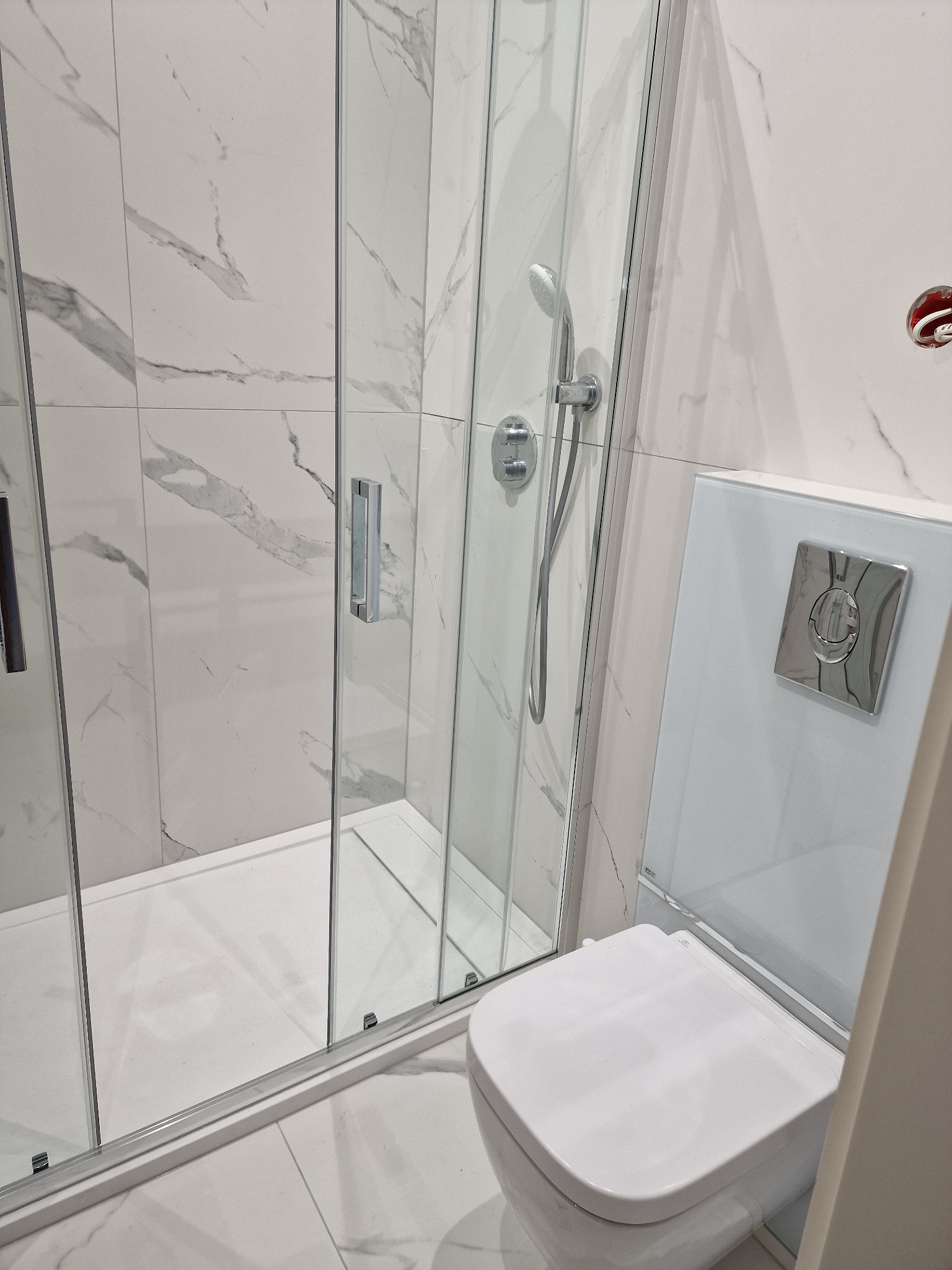 Nowoczesna łazienka z kabiną prysznicową ze szklanymi drzwiami i toaletą z białym panelem spłukującym na tle płytek imitujących marmur.