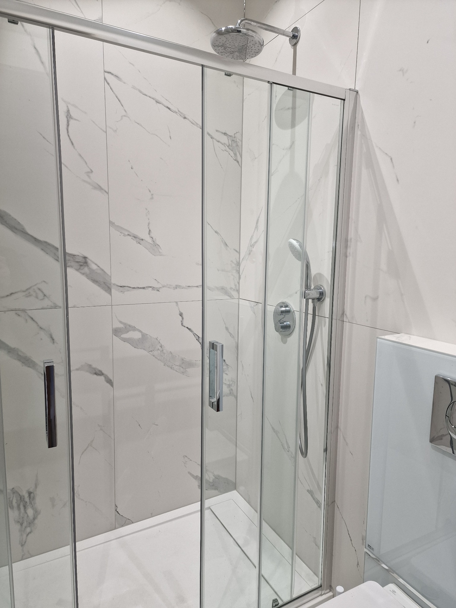 Nowoczesna kabina prysznicowa z przesuwnymi szklanymi drzwiami, wykończona białymi płytkami imitującymi marmur, z widoczną deszczownicą i słuchawką prysznicową.