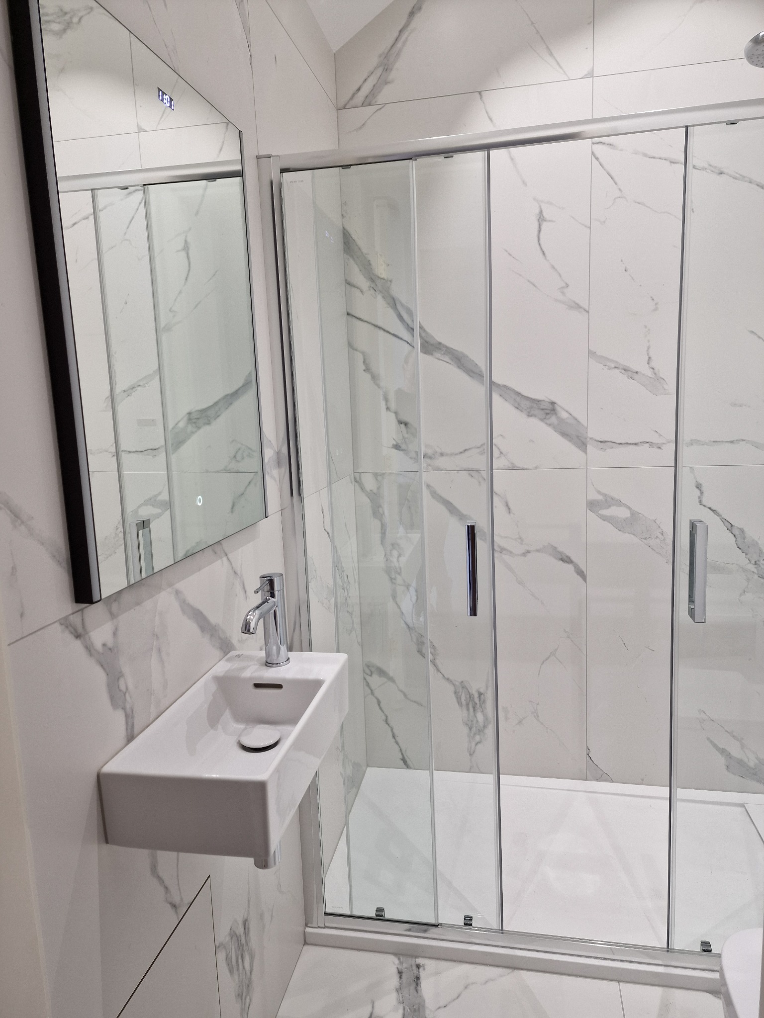Nowoczesna łazienka z białymi marmurowymi płytkami, kabiną prysznicową z przesuwnymi drzwiami i minimalistyczną umywalką z chromowaną baterią.