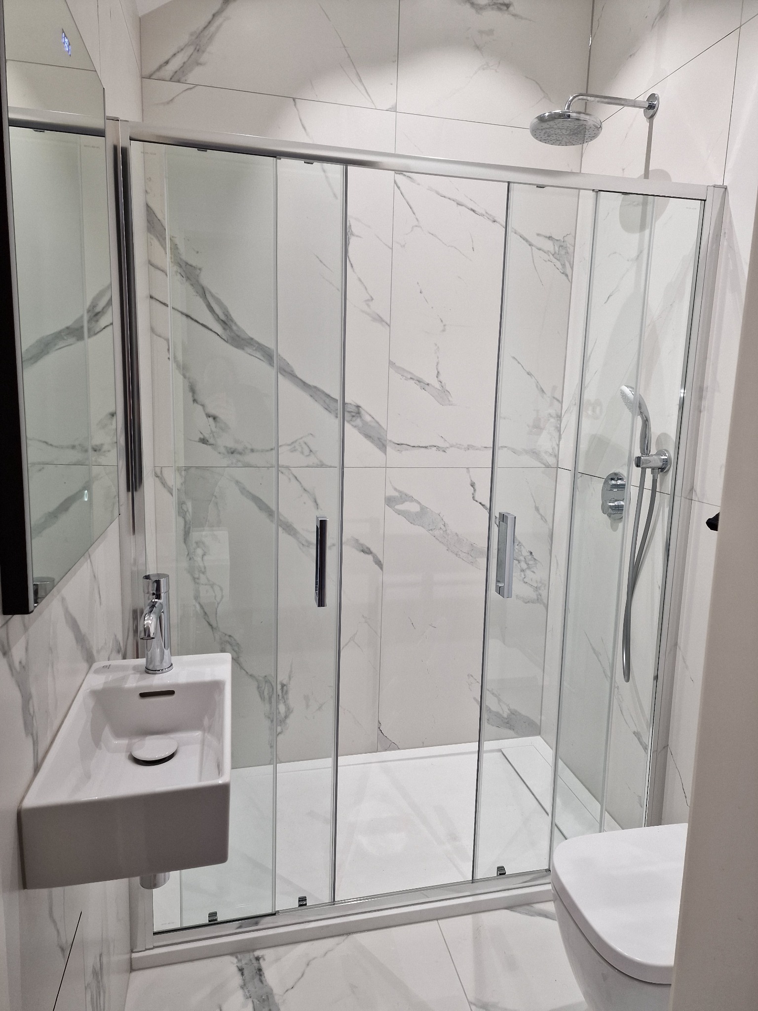 Nowoczesna łazienka z kabiną prysznicową z przesuwnymi drzwiami, umywalką nablatową i toaletą, wykończona białymi płytkami imitującymi marmur.