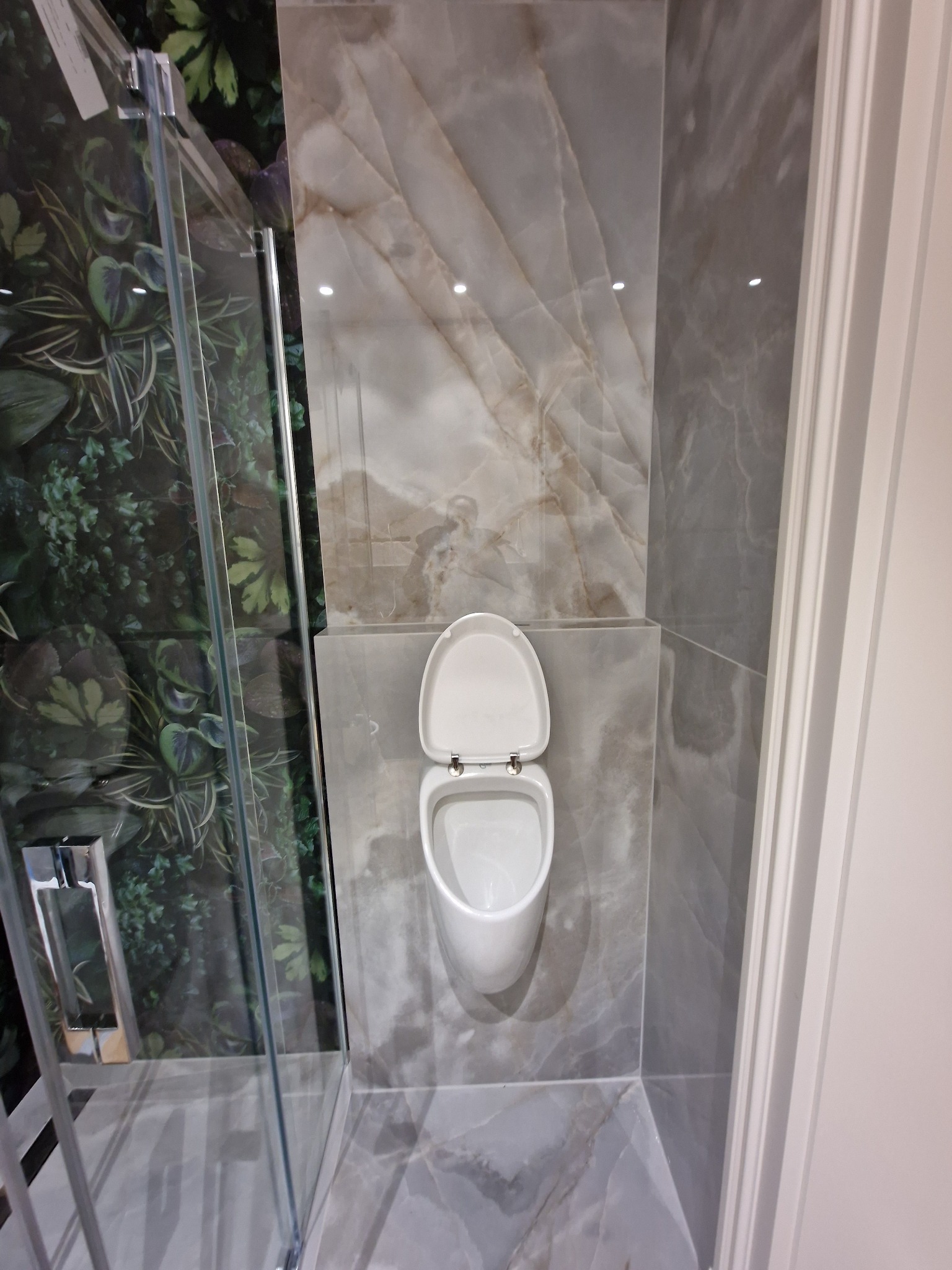Nowoczesna łazienka z wiszącą toaletą, marmurowymi płytkami i kabiną prysznicową z roślinnym motywem na ścianie.