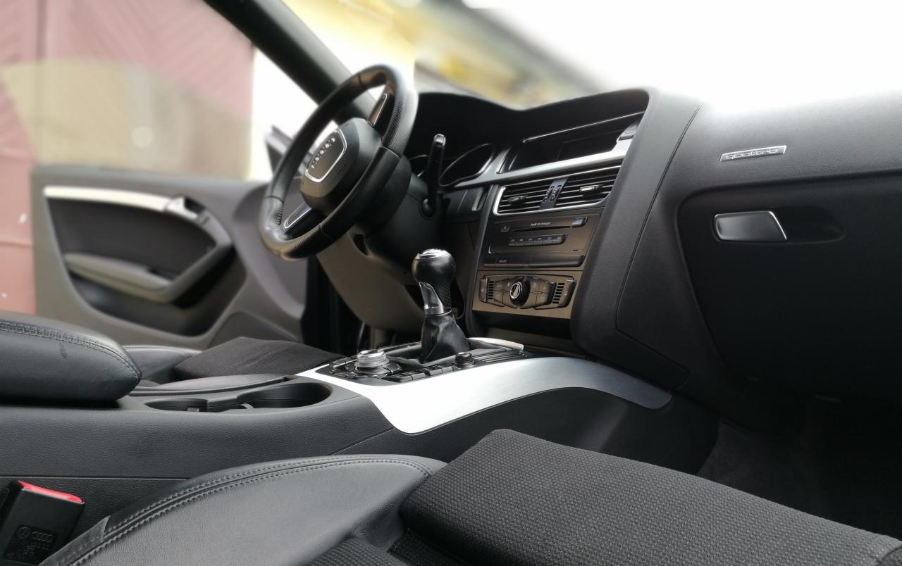 Wnętrze samochodu Audi A5 z czarną skórzaną tapicerką, srebrnymi akcentami na desce rozdzielczej i konsoli środkowej, widok z perspektywy pasażera na kierownicę i dźwignię zmiany biegów.