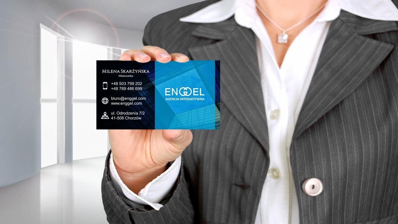 Osoba w garniturze prezentuje wizytówkę Agencji Interaktywnej ENGEL z danymi kontaktowymi Mileny Skarżyńskiej, właścicielki, na tle jasnego, minimalistycznego wnętrza biurowego.