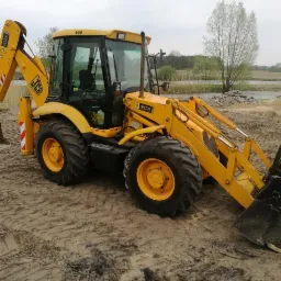 Żółta koparko-ładowarka JCB 3CX na piaszczystym terenie z widocznymi śladami opon, w tle drzewa i zbiornik wodny, pochmurne niebo.