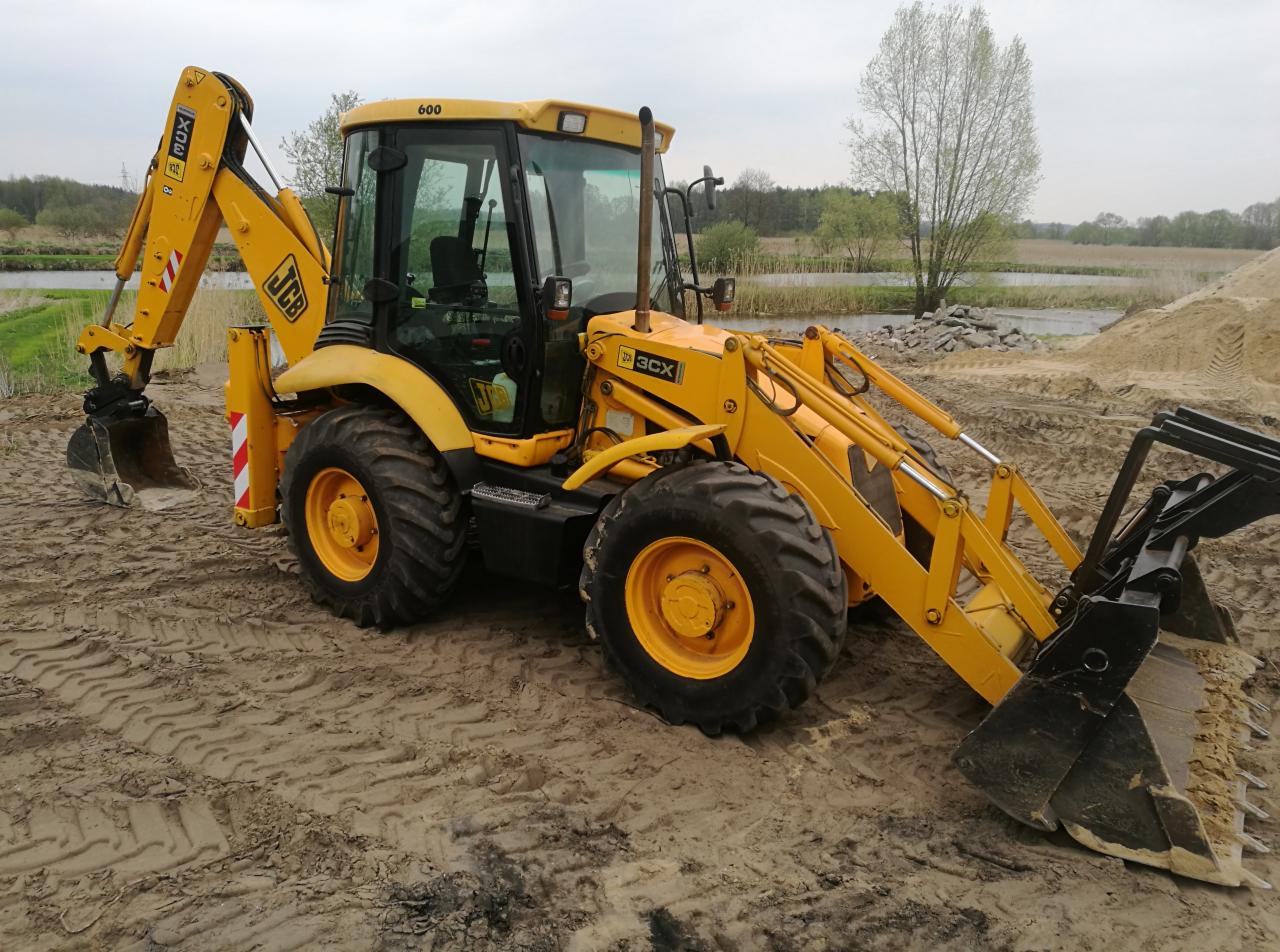 Żółta koparko-ładowarka JCB 3CX na piaszczystym terenie z widocznymi śladami opon, w tle drzewa i zbiornik wodny, pochmurne niebo.
