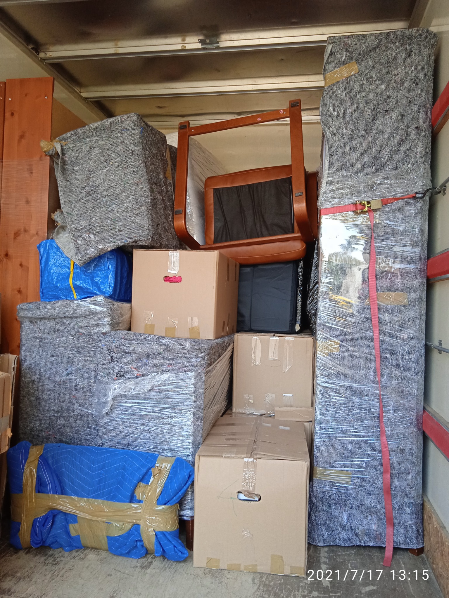 Wnętrze samochodu dostawczego załadowane kartonami, meblami owiniętymi folią i kocami transportowymi, zabezpieczone pasami.