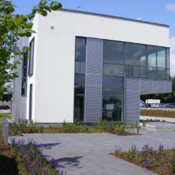 Nowoczesny, dwukondygnacyjny budynek biurowy z białymi ścianami i perforowanymi panelami elewacyjnymi, otoczony kostką brukową i rabatami z lawendy, widok z poziomu chodnika w słoneczny dzień.