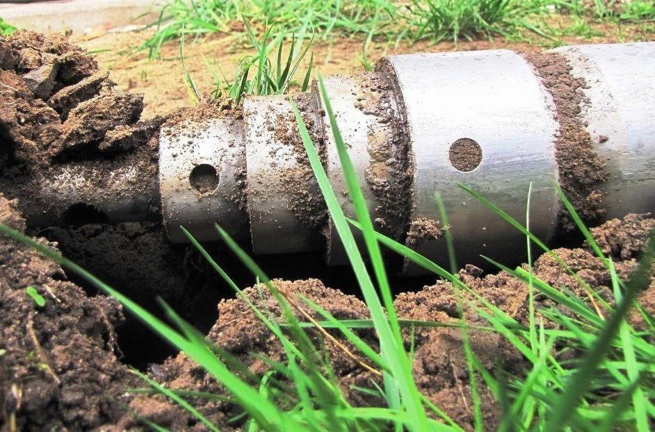 Metalowa rura z otworami, częściowo zagłębiona w ziemi, otoczona zieloną trawą, widoczne ślady ziemi na powierzchni rury.