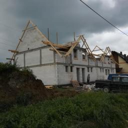 Karpier general construction - Wznoszenie dwukondygnacyjnego domu z betonu komórkowego, widoczne drewniane elementy konstrukcji dachu i pracownik w oddali, pochmurne niebo w tle.