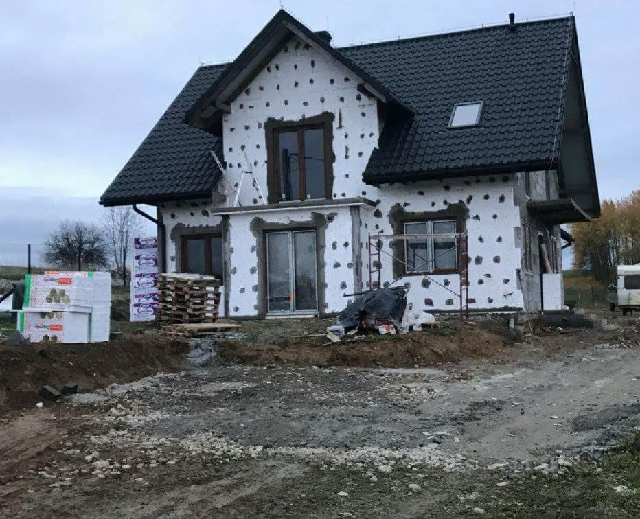 Dom w trakcie ocieplania styropianem, widoczne rusztowania, palety z materiałami budowlanymi i ziemia wokół budynku.