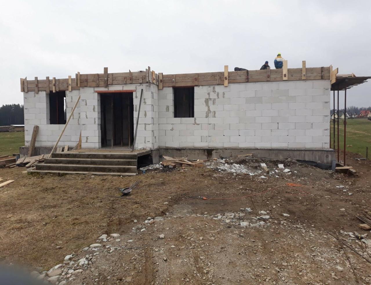 Budowa domu jednorodzinnego w stanie surowym otwartym, ściany z białych bloczków, widoczne szalunki na stropie i pracownicy na górze.