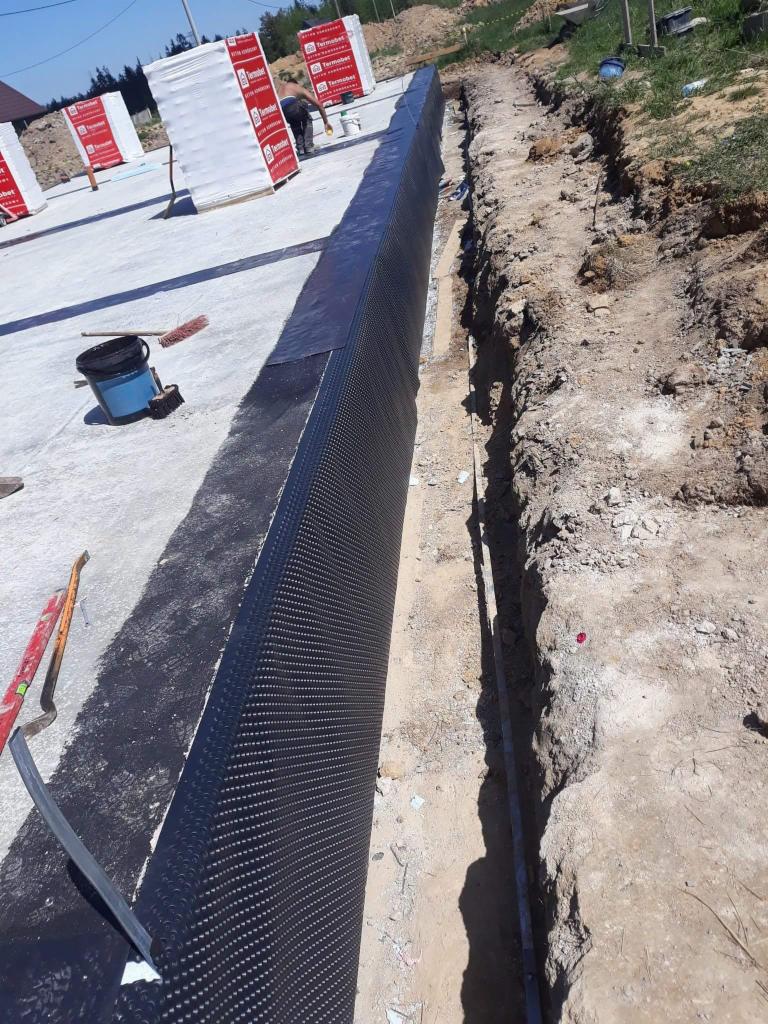 Izolacja fundamentu budynku w trakcie budowy, czarna folia kubełkowa zabezpiecza ścianę fundamentową, wykop ziemny obok, widoczne materiały budowlane i narzędzia.