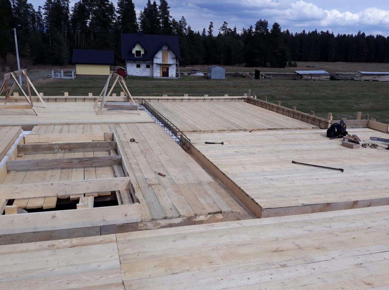 Drewniany szalunek pod płytę fundamentową z widocznym zbrojeniem i narzędziami, w tle dom w trakcie budowy.