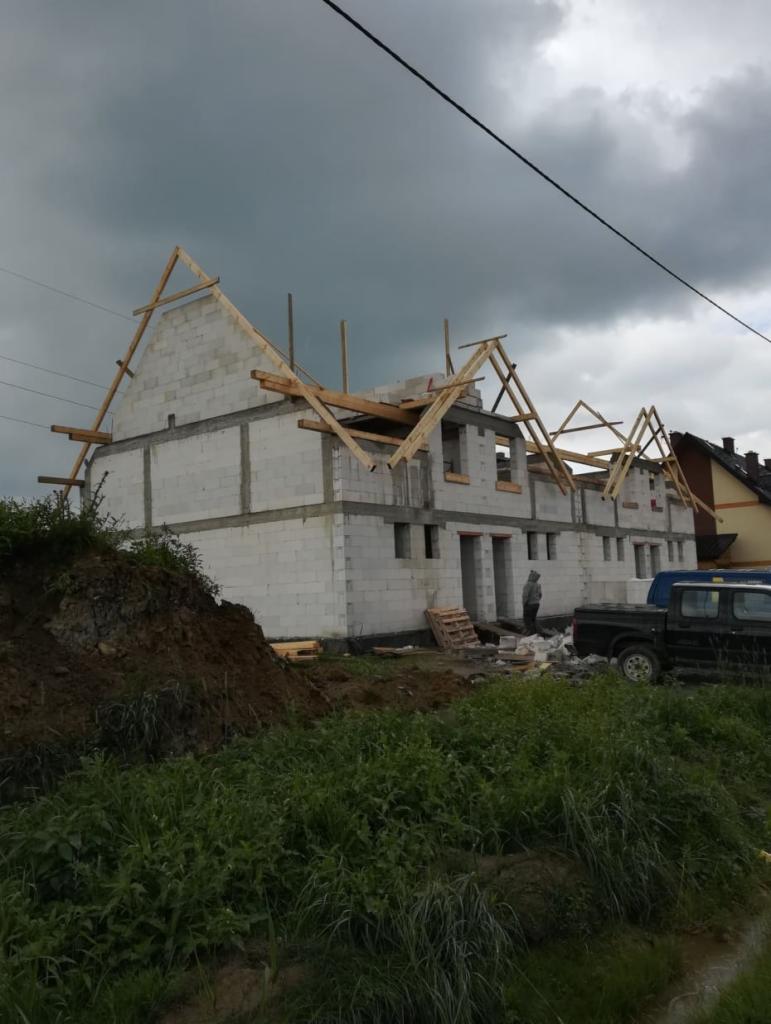 Wznoszenie dwukondygnacyjnego domu z betonu komórkowego, widoczne drewniane elementy konstrukcji dachu i pracownik w oddali, pochmurne niebo w tle.