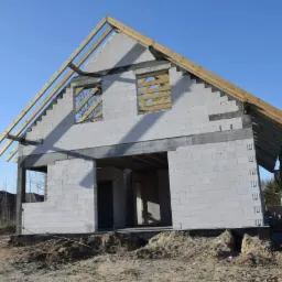 Surowy stan domu murowanego z bloczków betonowych, widoczne drewniane krokwie dachu, niezabezpieczone otwory okienne i nieuregulowany teren wokół budynku.