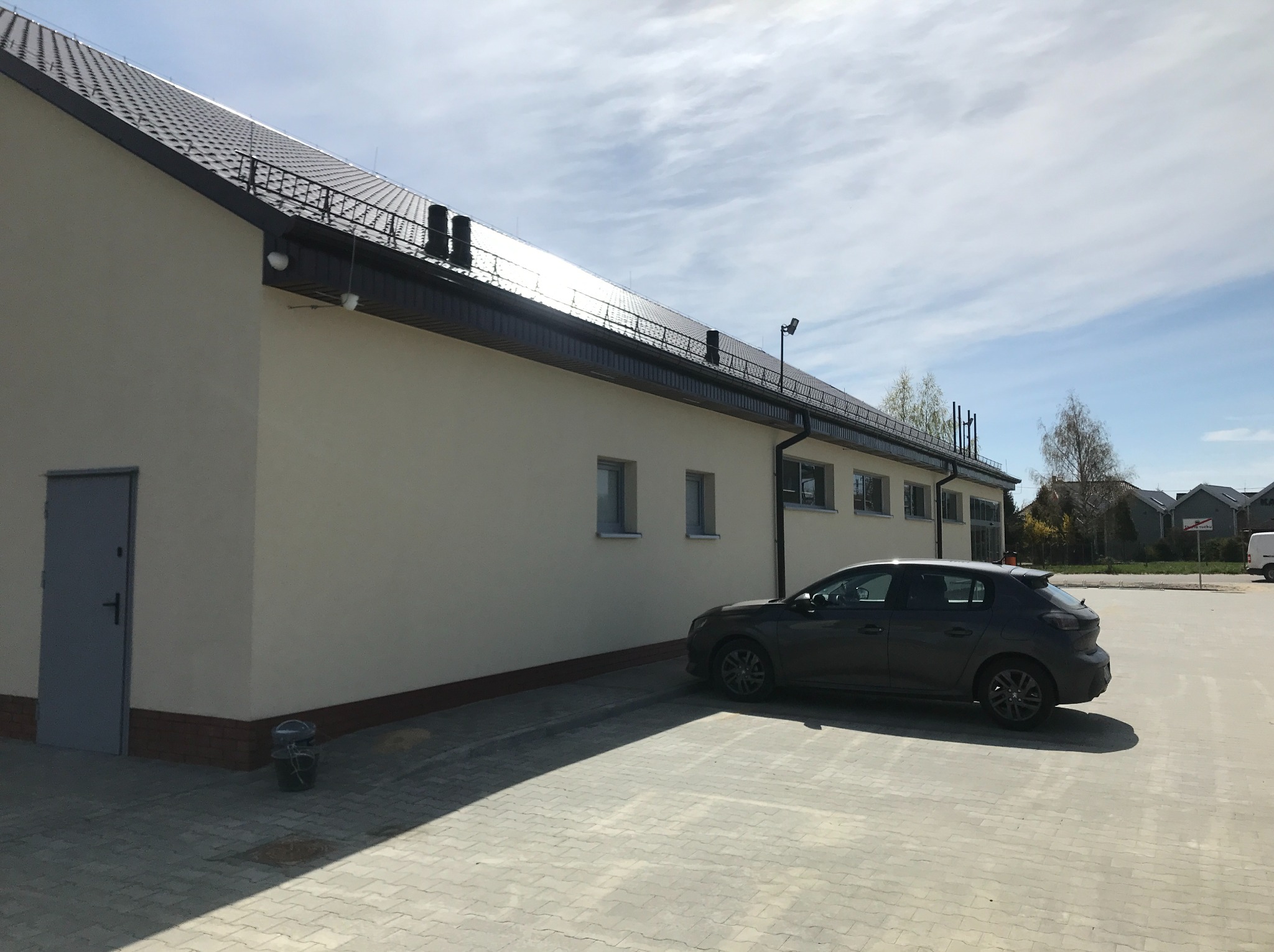 Elewacja budynku z ciemną dachówką i rynnami, zaparkowany szary samochód osobowy. Widok na fragment budynku i otoczenie w słoneczny dzień.