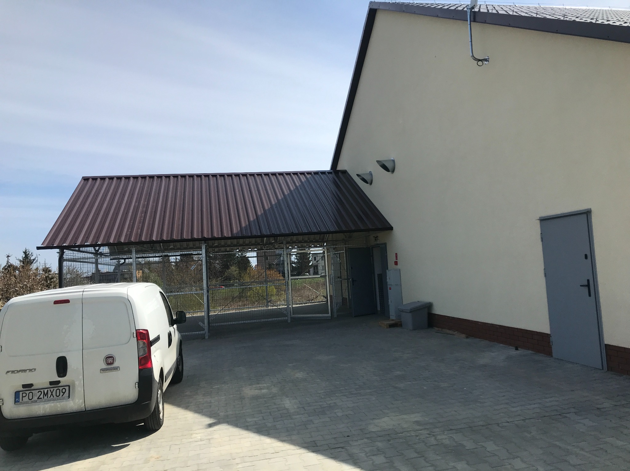 Jasny budynek z brązowym dachem, szarą bramą i białym samochodem dostawczym zaparkowanym przed nim, w słoneczny dzień. Widoczny fragment elewacji.