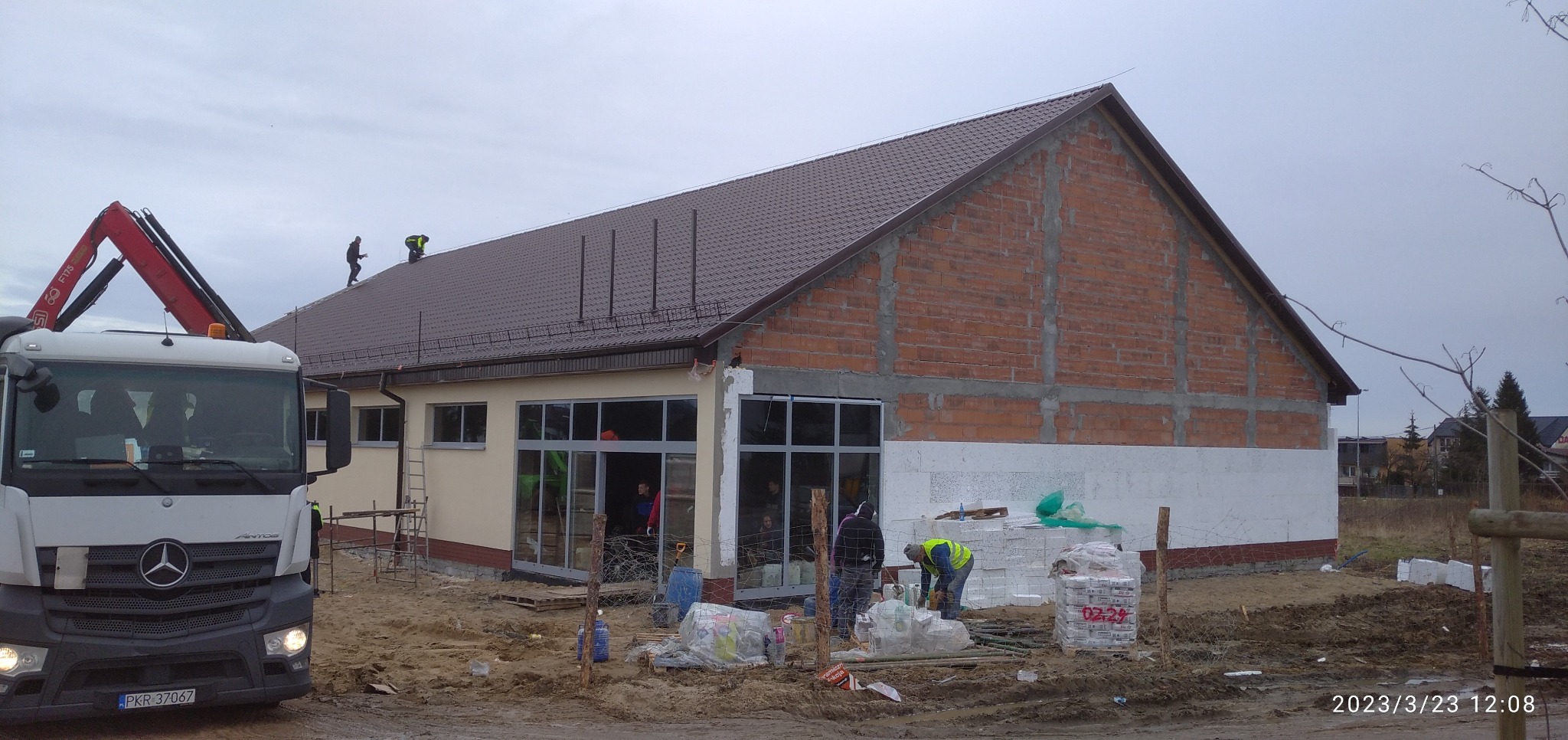 Budowa hali w Gdańsku: ekipa na dachu, ocieplanie ścian styropianem, okna wstawione, ceglany szczyt. Widoczny dźwig i materiały budowlane.